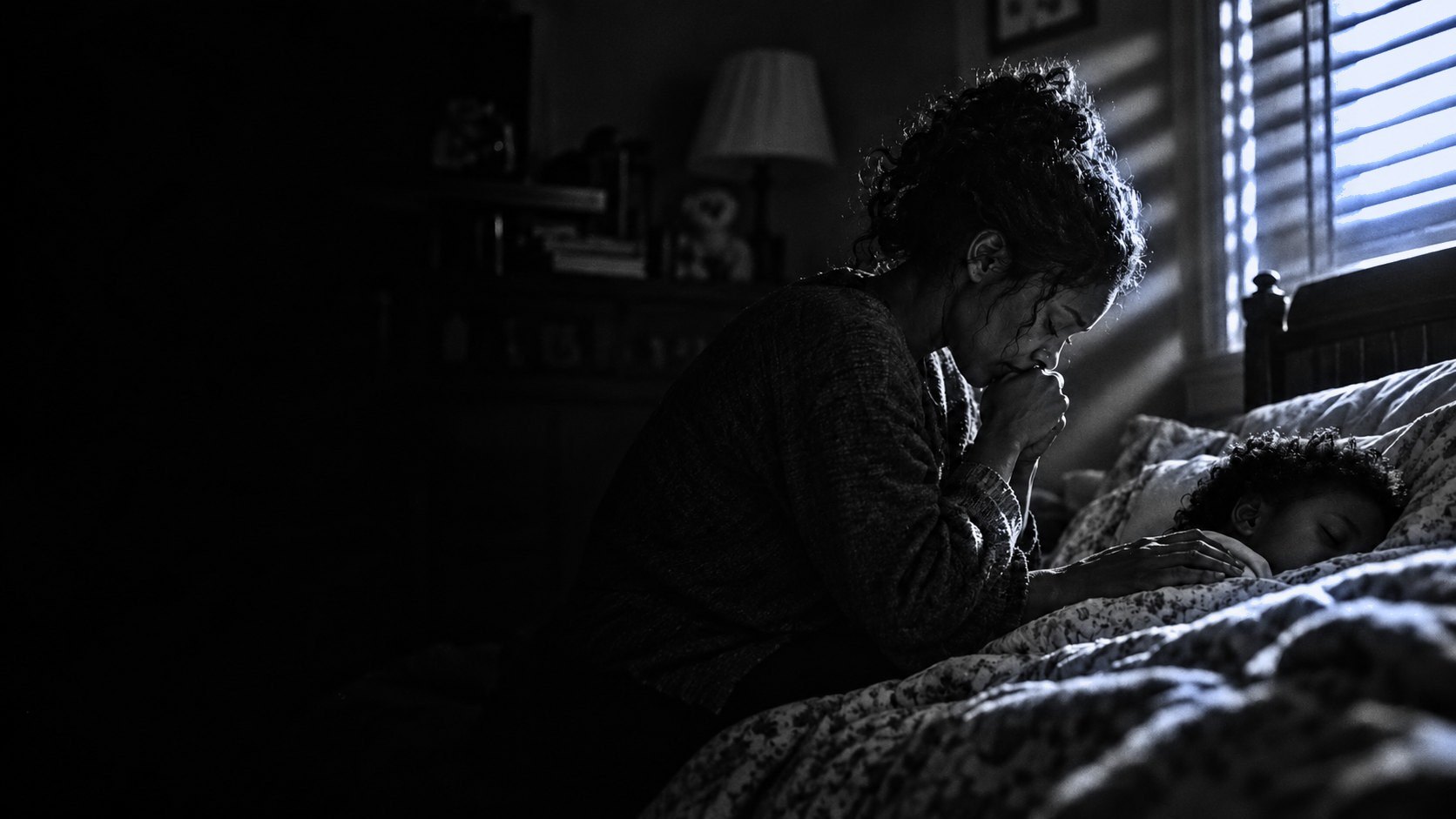 A young man in a blue hoodie kneeling beside his bed at sunrise, head bowed into his hands in prayer, his Bible open beside him